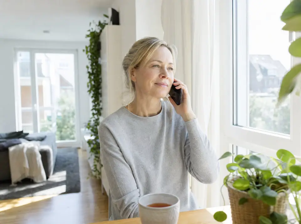 Eine Frau sitzt in einer hellen Wohnung am Fenster, telefoniert entspannt und hat eine Tasse Tee vor sich.